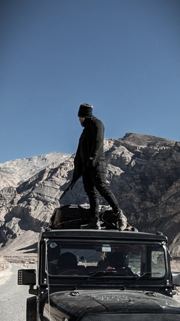 a man standing on a SUV looking back at the mountains behind